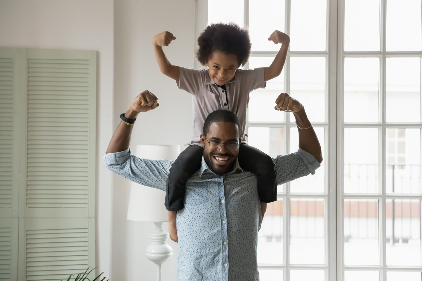 A father flexing his muscles with his child sitting on his shoulders, flexing